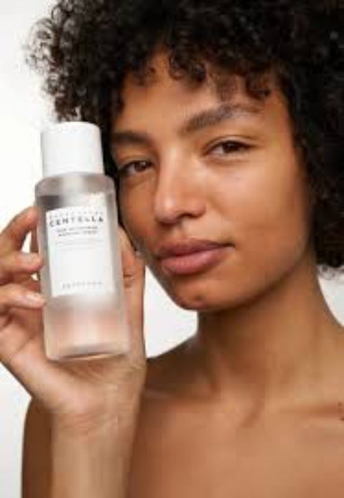 Young woman with curly hair holding SKIN1004 Centella toner bottle close to her face, smiling softly.